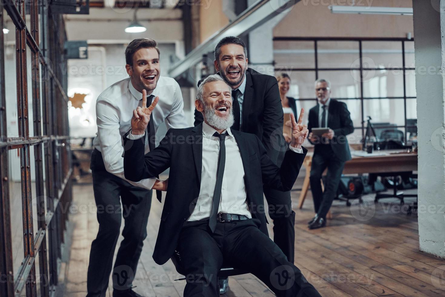 Cheerful businessmen pushing their boss on the office chair while running in the hallway photo