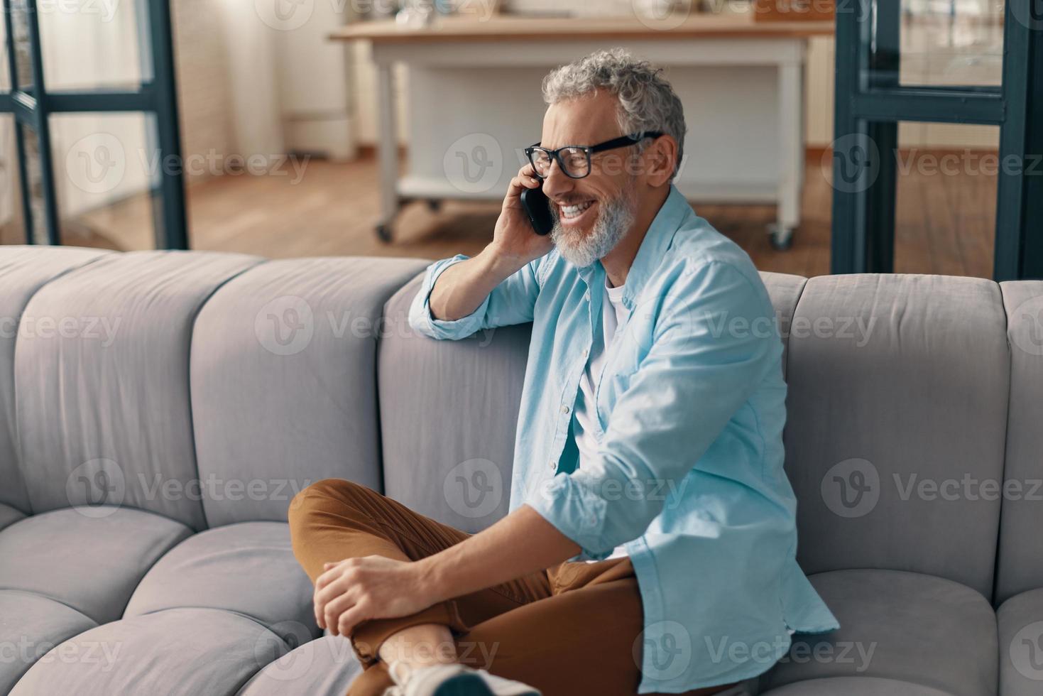 Happy senior man in casual clothing and eyeglasses talking on the smart phone while sitting on the sofa at home photo