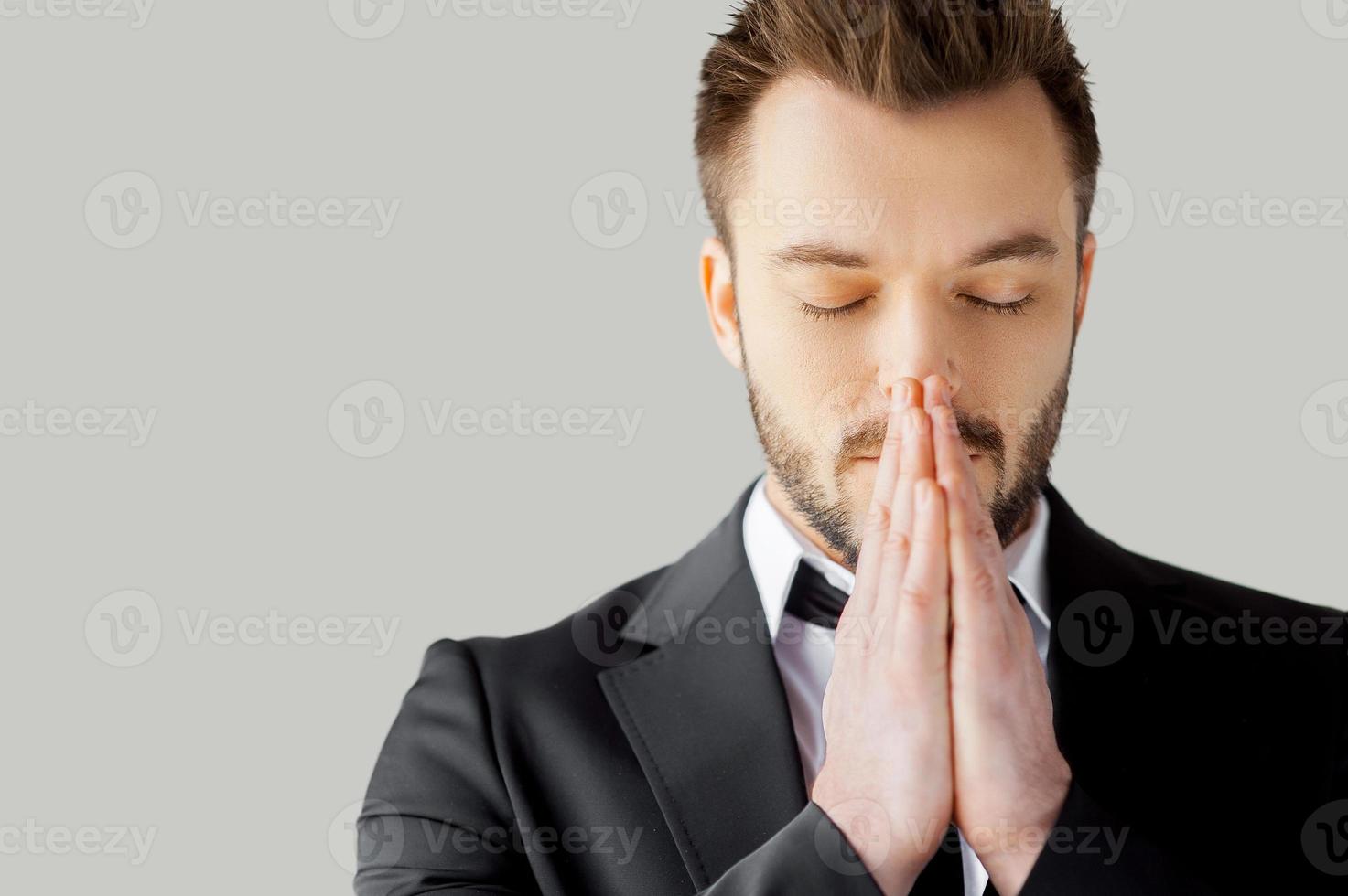Businessman praying. Portrait of young man in formalwear holding hands clasped and keeping eyes closed while standing against grey background photo