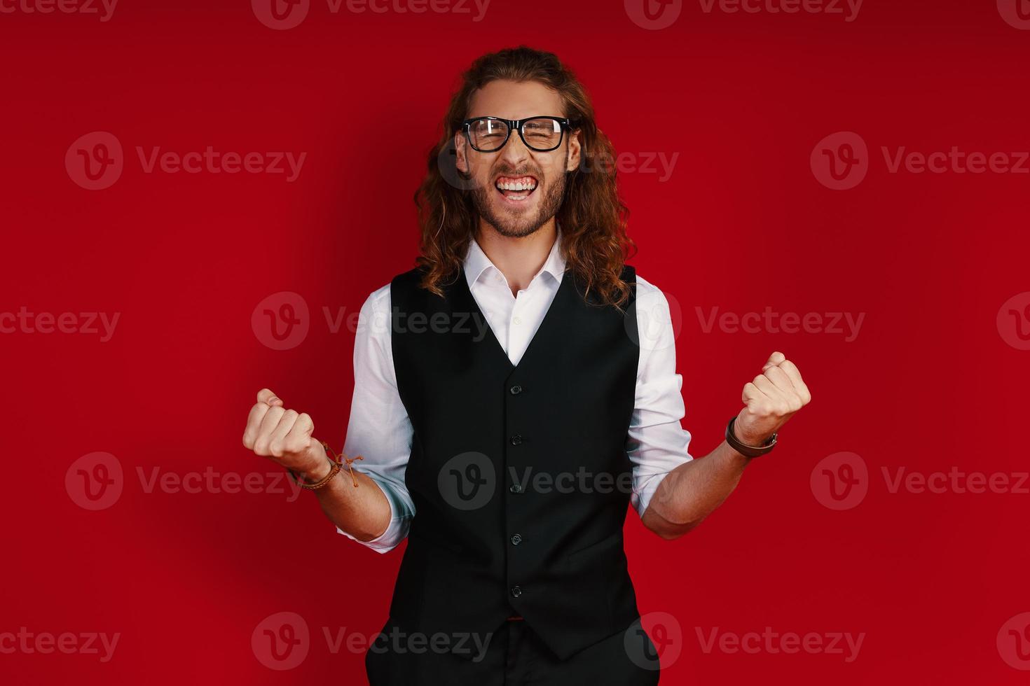 joven feliz con ropa elegante animando y gesticulando mientras está de pie contra el fondo rojo foto