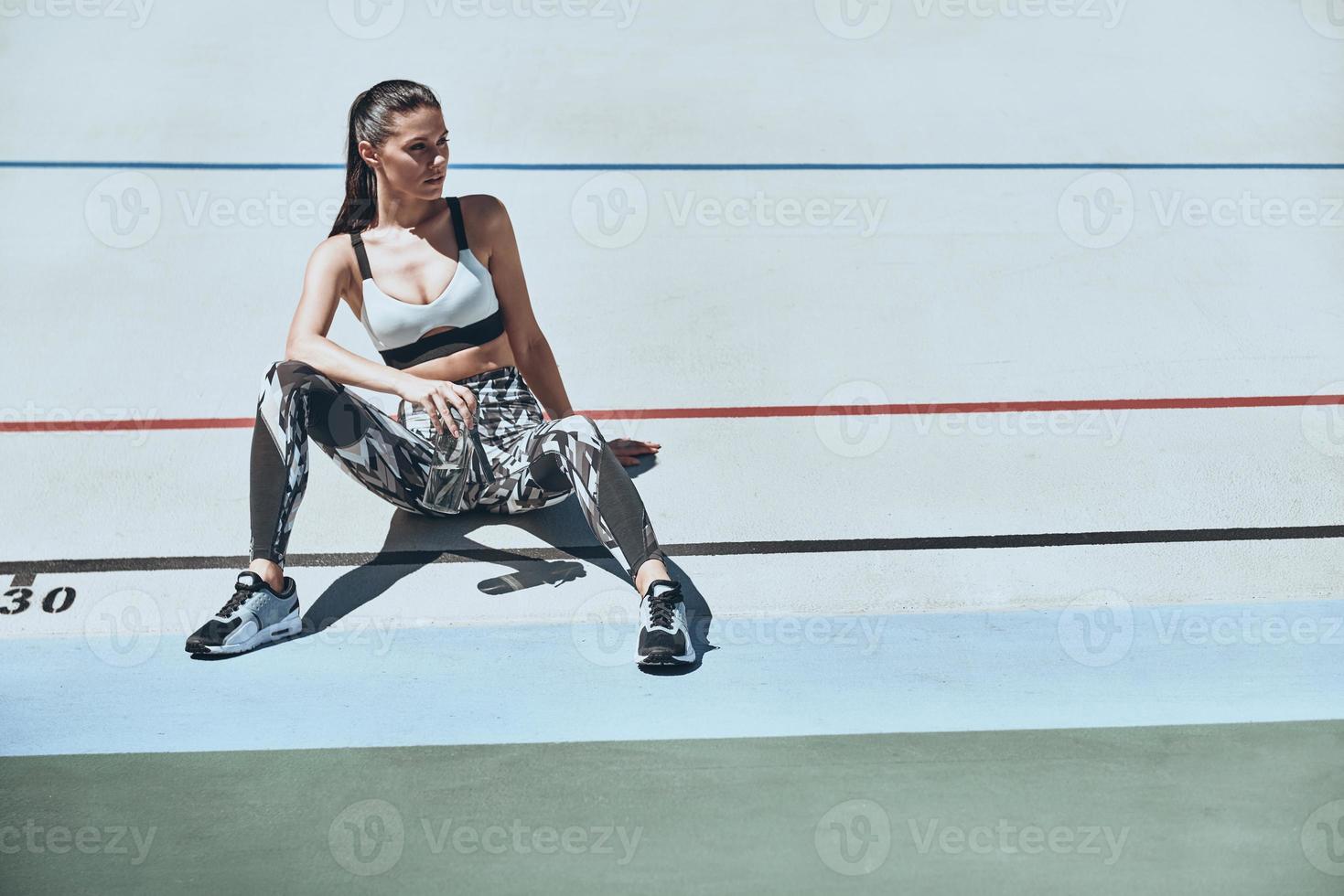 Taking a timeout. Beautiful young woman in sports clothing looking away while sitting on the running track outdoors photo