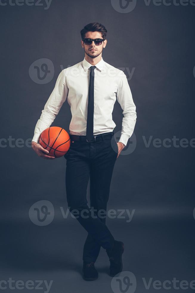 listo para jugar toda la longitud de un joven apuesto y bien vestido con gafas de sol sosteniendo una pelota de baloncesto y mirando la cámara mientras se enfrenta a un fondo gris foto