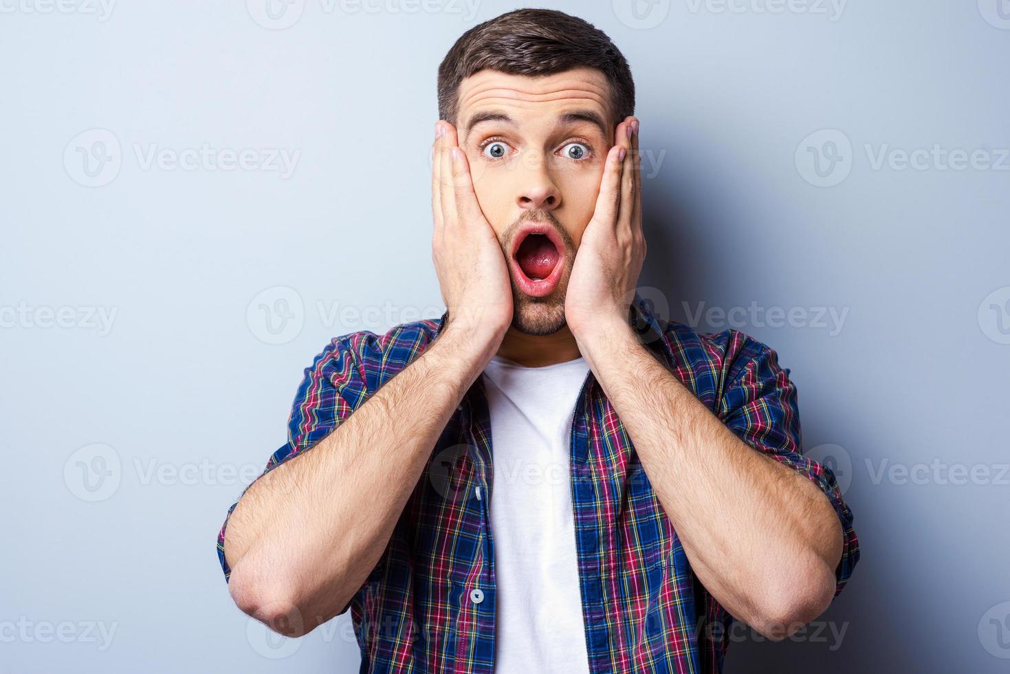 Feeling shocked. Frustrated young man in casual shirt keeping mouth open and looking terrified while touching his face and standing against grey background photo