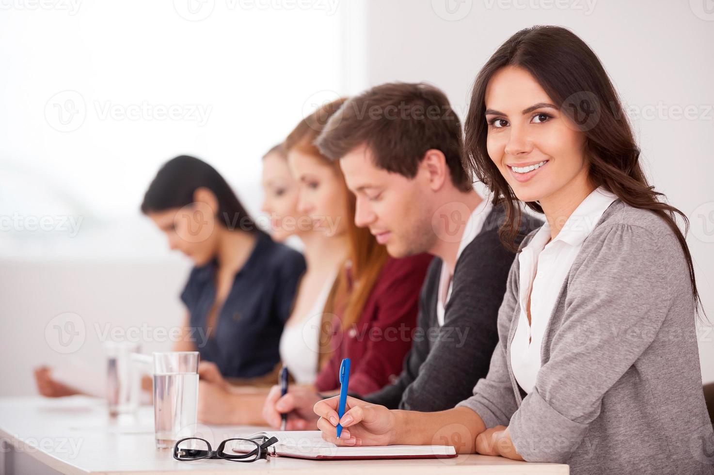 gente en el seminario. atractiva mujer joven sonriendo a la cámara mientras se sienta junto a otra gente en la mesa foto