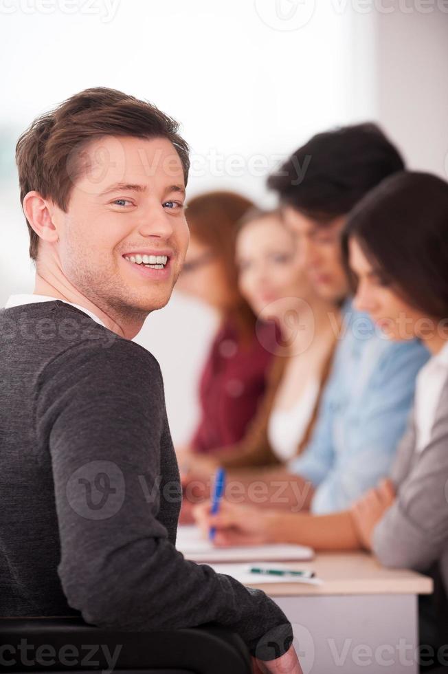 Confident in his team. Rear view of handsome young man looking over shoulder and smiling while sitting together at the table with another people photo