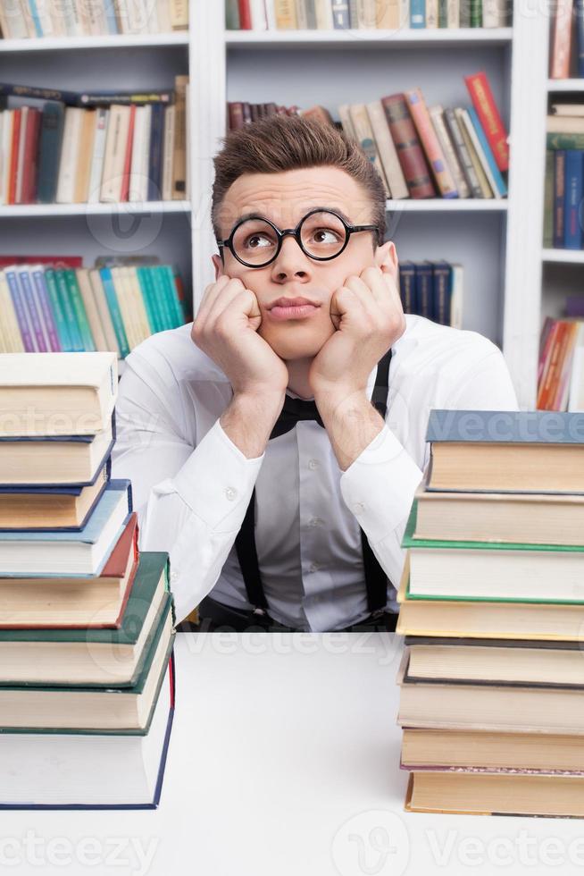 Nerd dreaming. Bored young man in shirt and bow tie sitting at the table in library and holding head in hands while two book stacks laying on the foreground photo