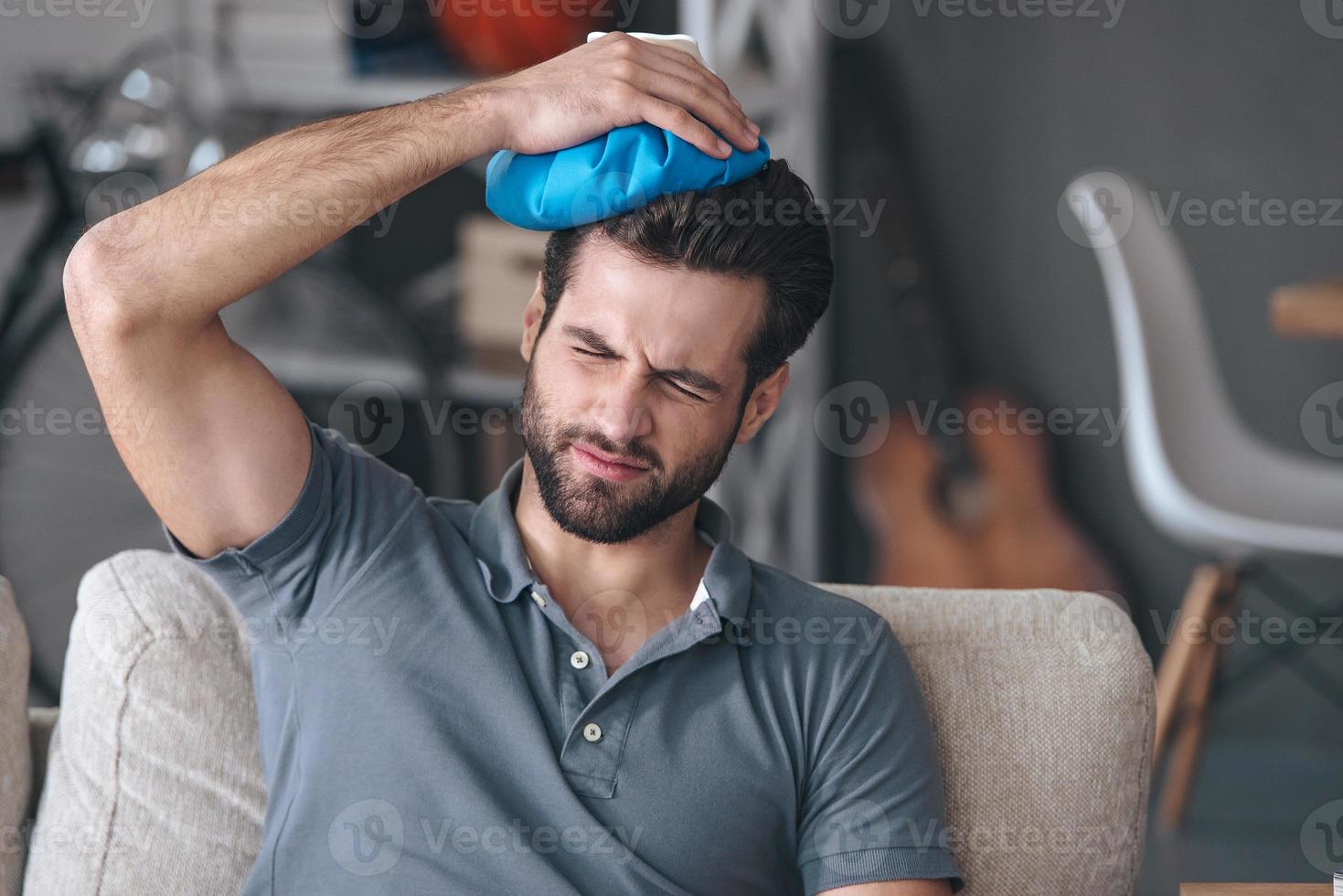 Headache that kills him. Frustrated handsome young man holding ice bag on his head while sitting on the couch at home photo