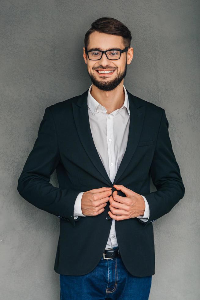 Confident smile. Cheerful young man in glasses looking at camera with smile and buttoning his jacket while standing against grey background photo