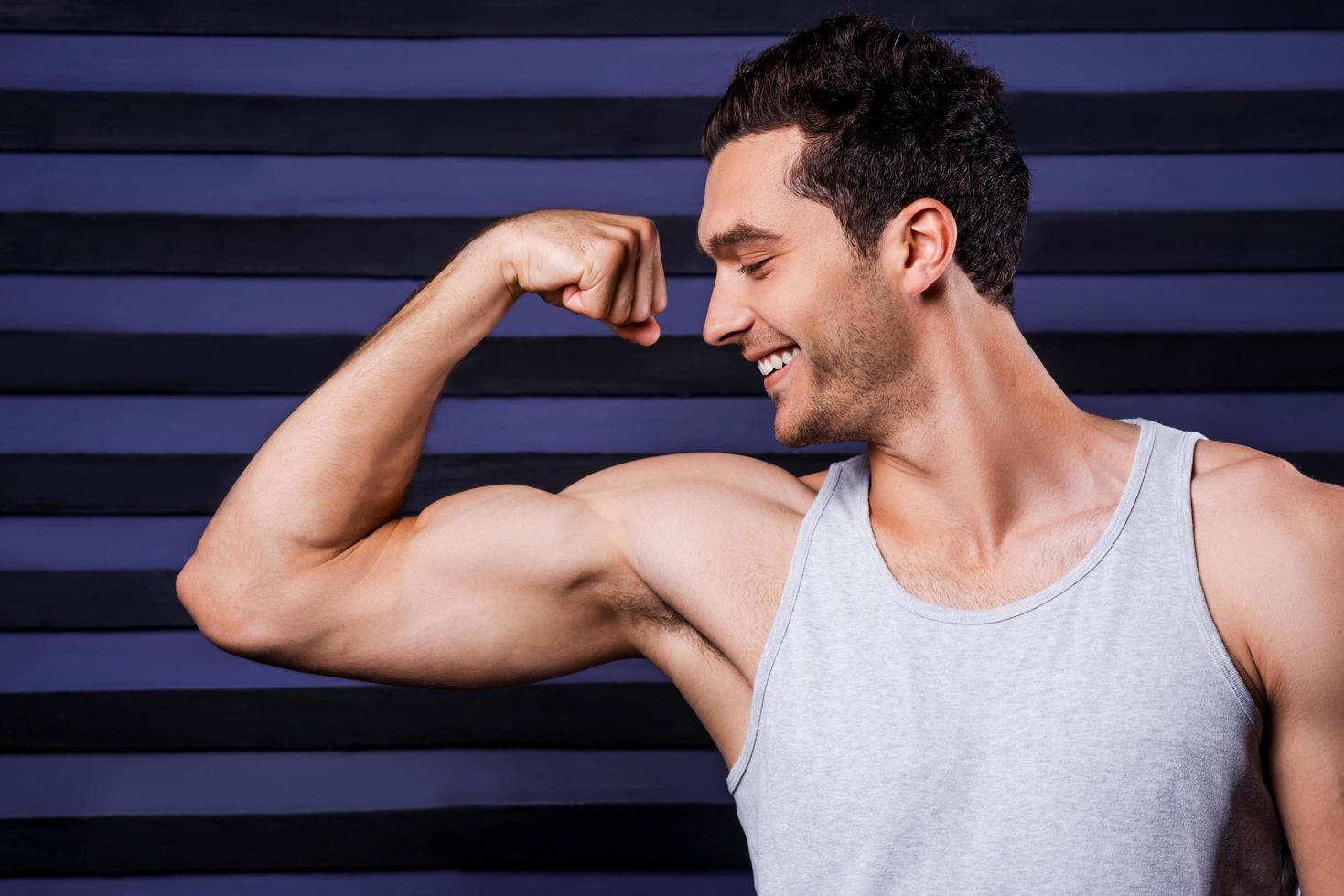 tiene un cuerpo perfecto. joven musculoso en la parte superior del tanque examinando sus bíceps perfectos y sonriendo mientras está de pie contra el fondo rayado foto