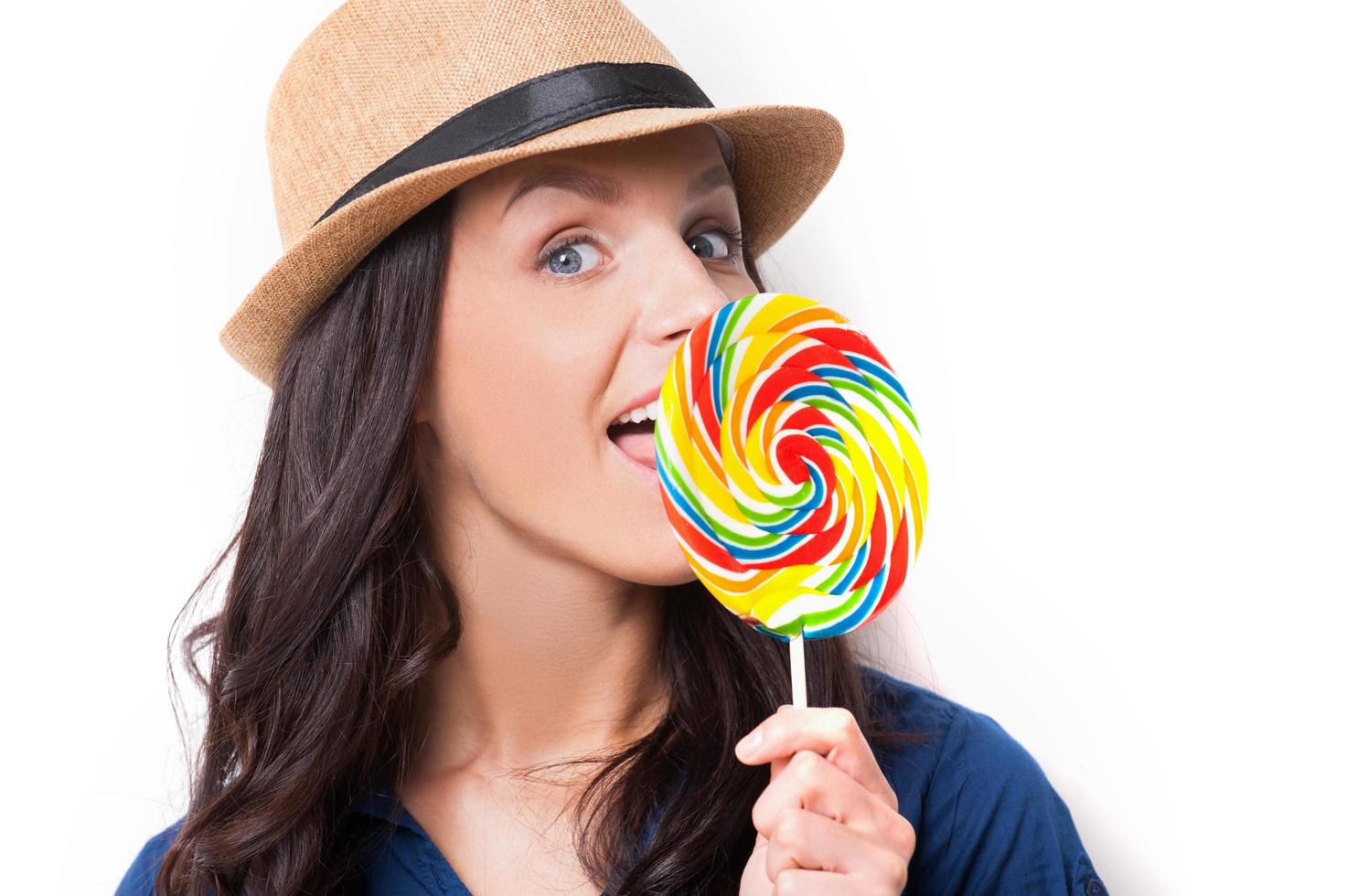 Sweet and extremely big. Surprised young beautiful woman in funky hat licking a big lollipop while standing against white background photo