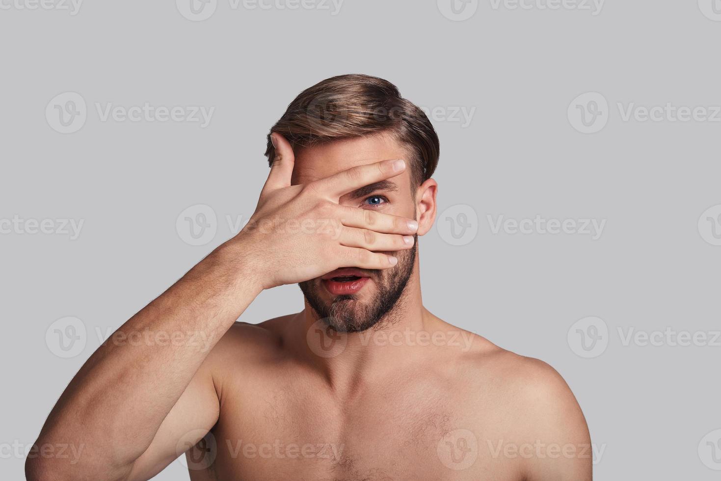 I want to know everything. Handsome young man covering eye with hand and peeking while standing against grey background photo