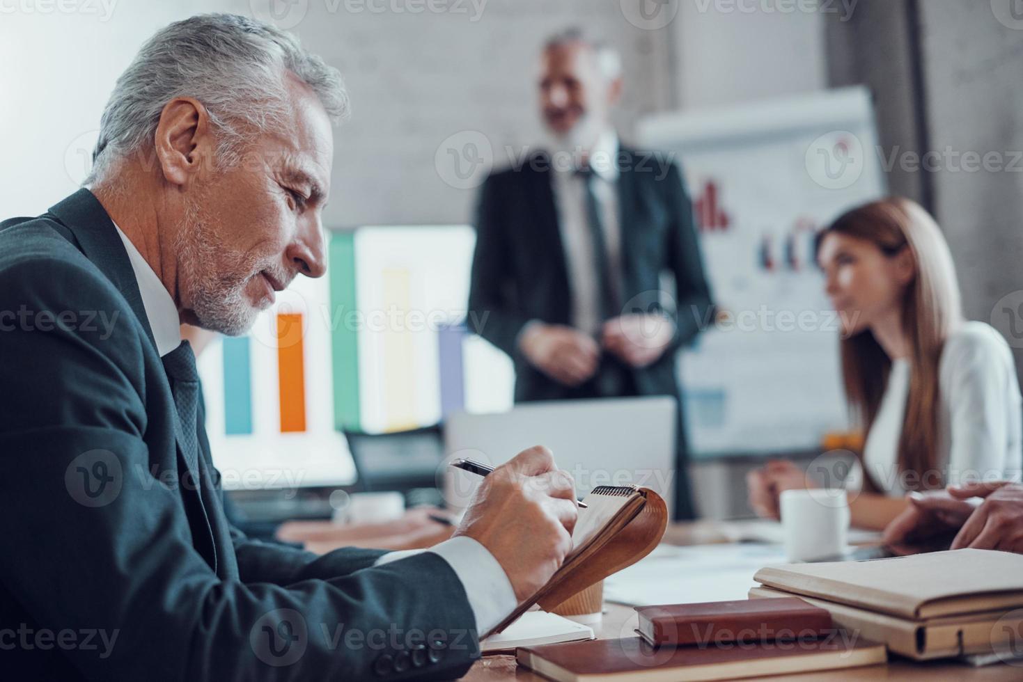 hombre de negocios maduro escribiendo algo y sonriendo mientras trabaja junto con colegas en la sala de juntas foto