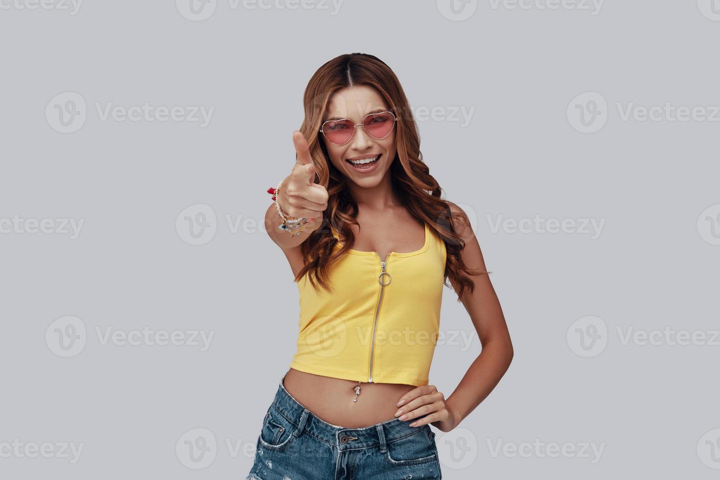 Attractive young woman looking at camera and pointing you while standing against grey background photo