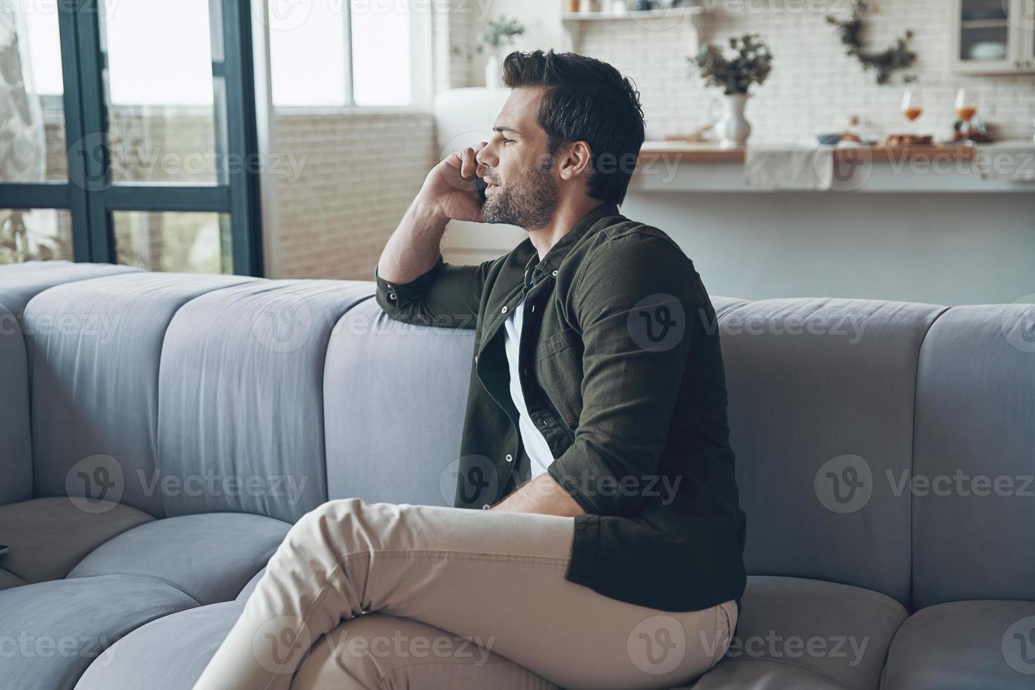 Handsome young man talking on smart phone and looking away while sitting on the sofa photo