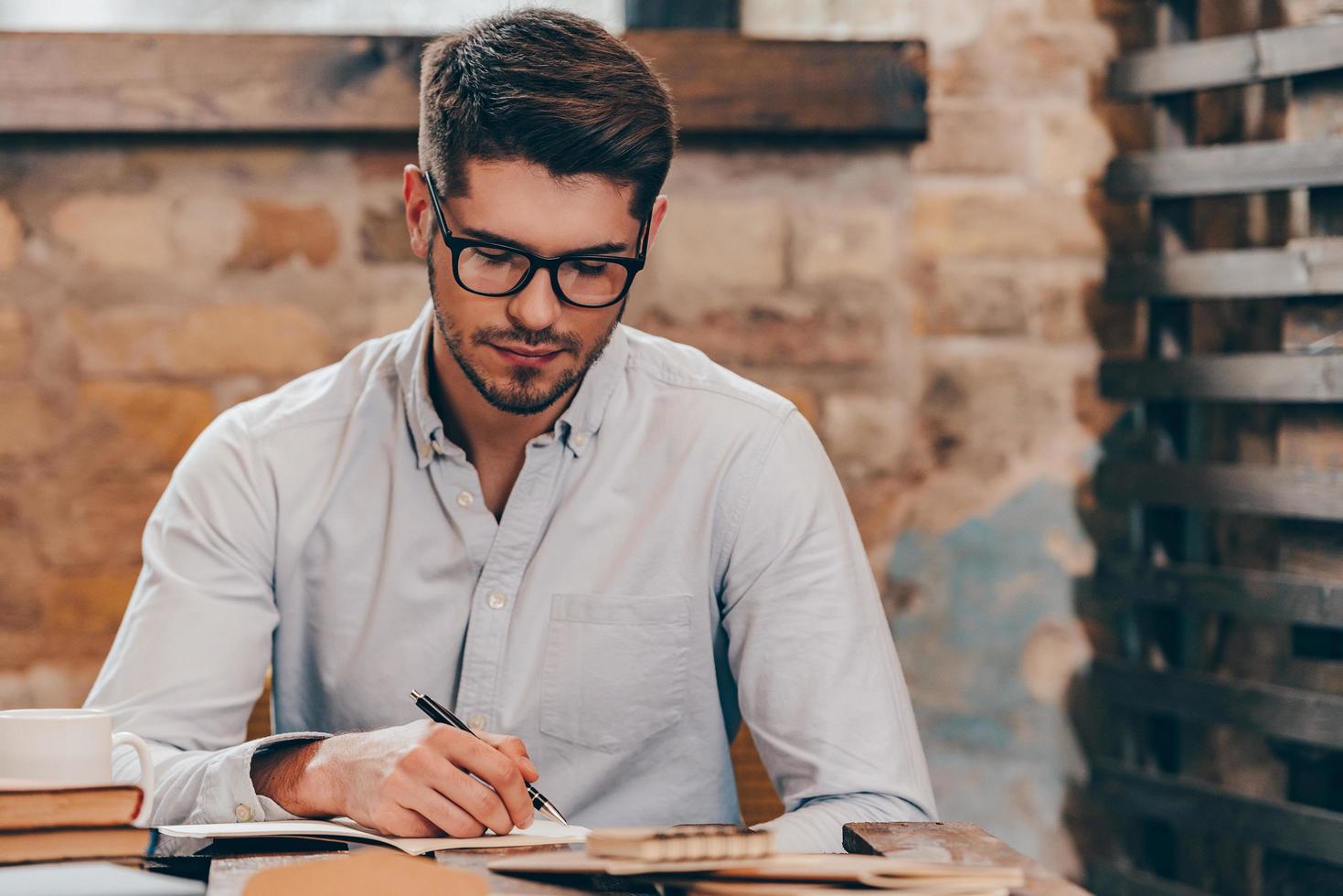 Making some notes. Handsome young man in glasses writing in his note pad while sitting at his working place photo