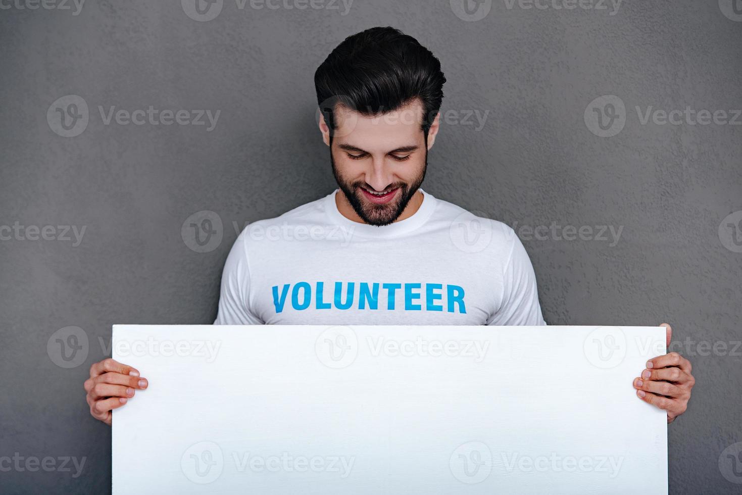 Please help to community Confident young man in volunteer t-shirt holding white board and looking at it with smile while standing against grey background photo