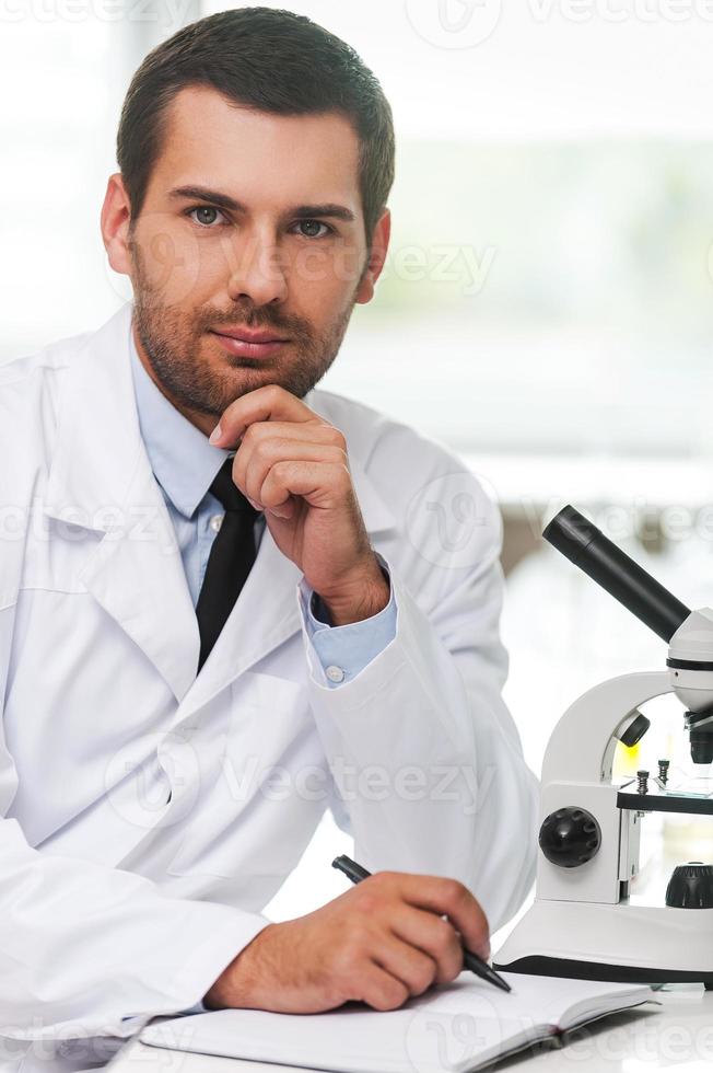 Confident scientist. Handsome young scientist in white uniform holding hand on chin and writing in note pad while sitting at his working place photo