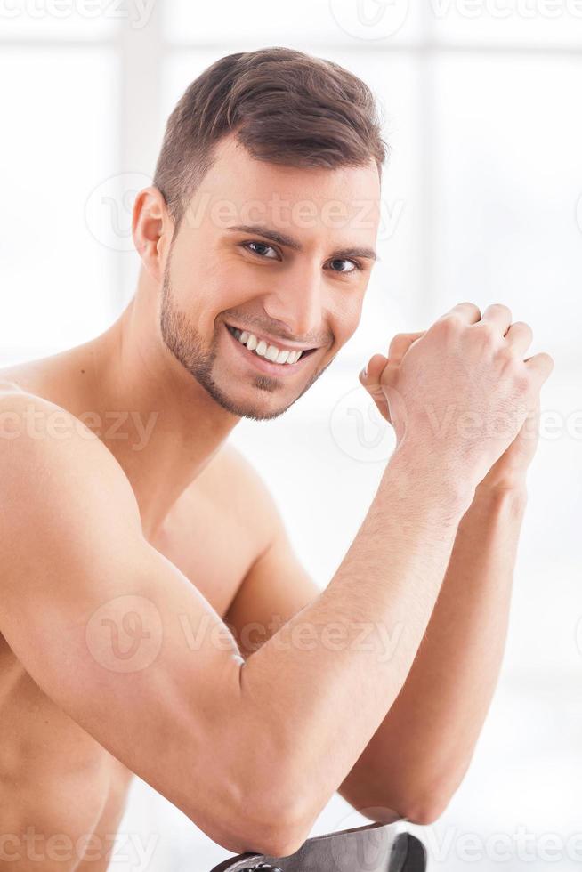 Feeling confident. Side view of handsome young muscular man sitting on the chair and smiling at camera photo