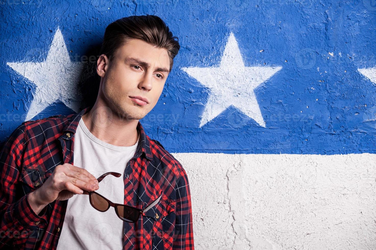 Thoughtful handsome. Handsome male model in checked shirt holding sunglasses while standing against background photo