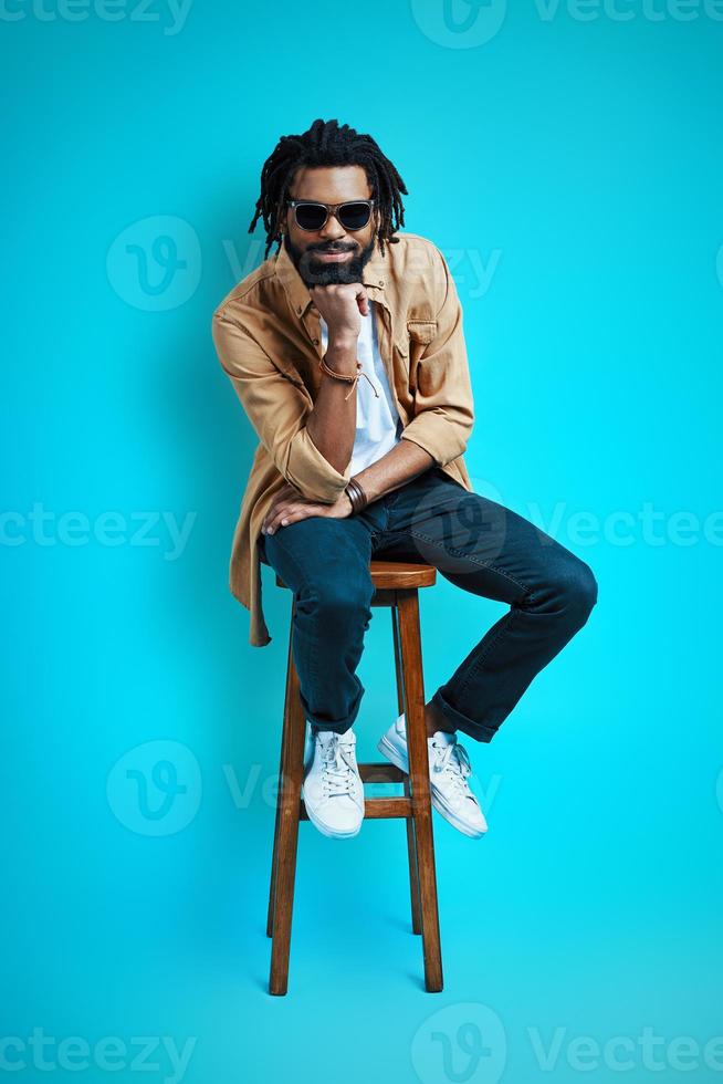 Full length of handsome young African man in casual wear looking at camera and smiling while sitting on stool against blue background photo