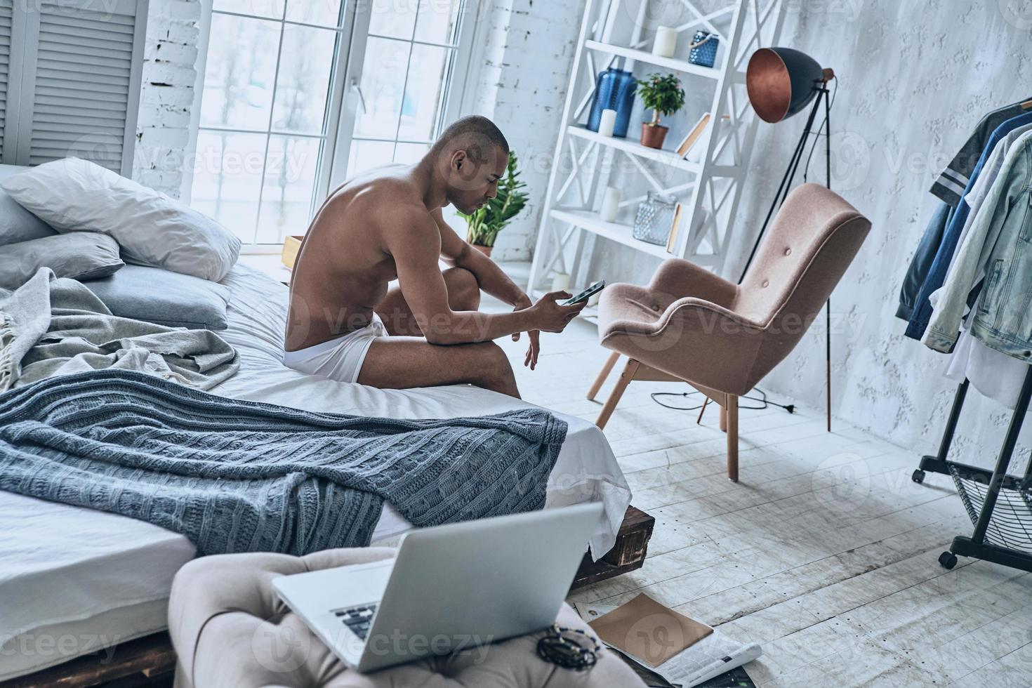 Chatting with girlfriend. Sleepy young African man using his smart phone while sitting on the bed at home photo