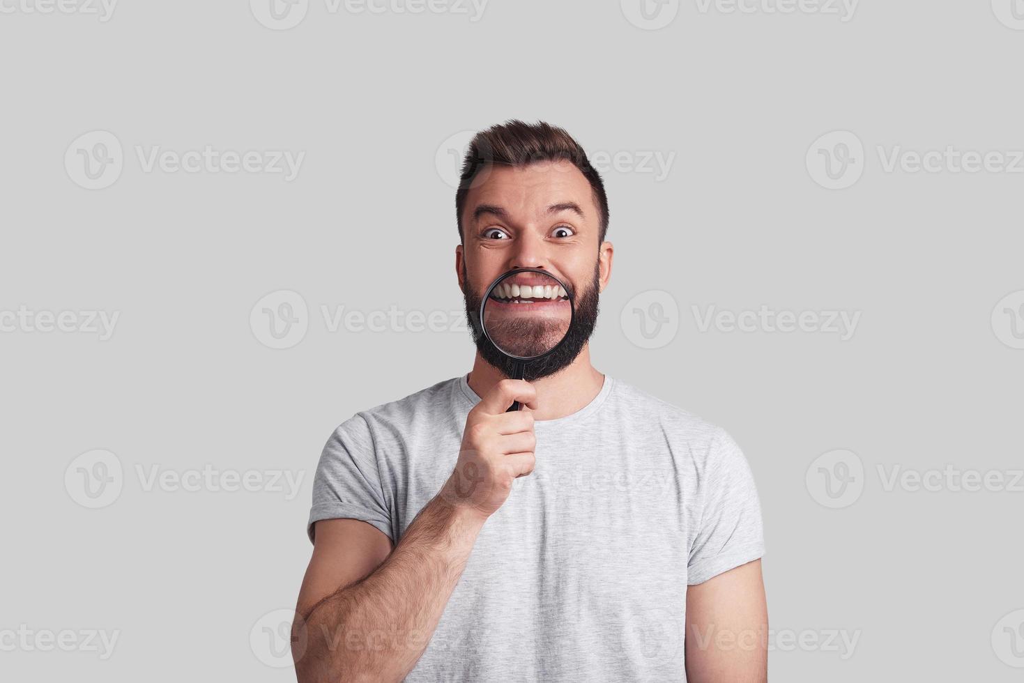 Perfect smile. Playful young man applying magnifying glass and smiling while standing against grey background photo
