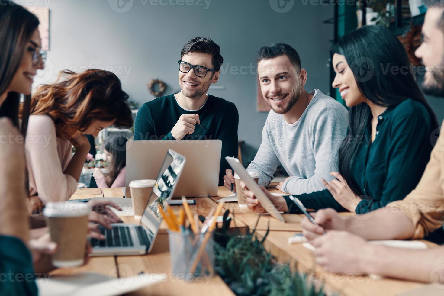 Putting ideas into something real. Group of young modern people in smart casual wear discussing something and smiling while working in the creative office photo