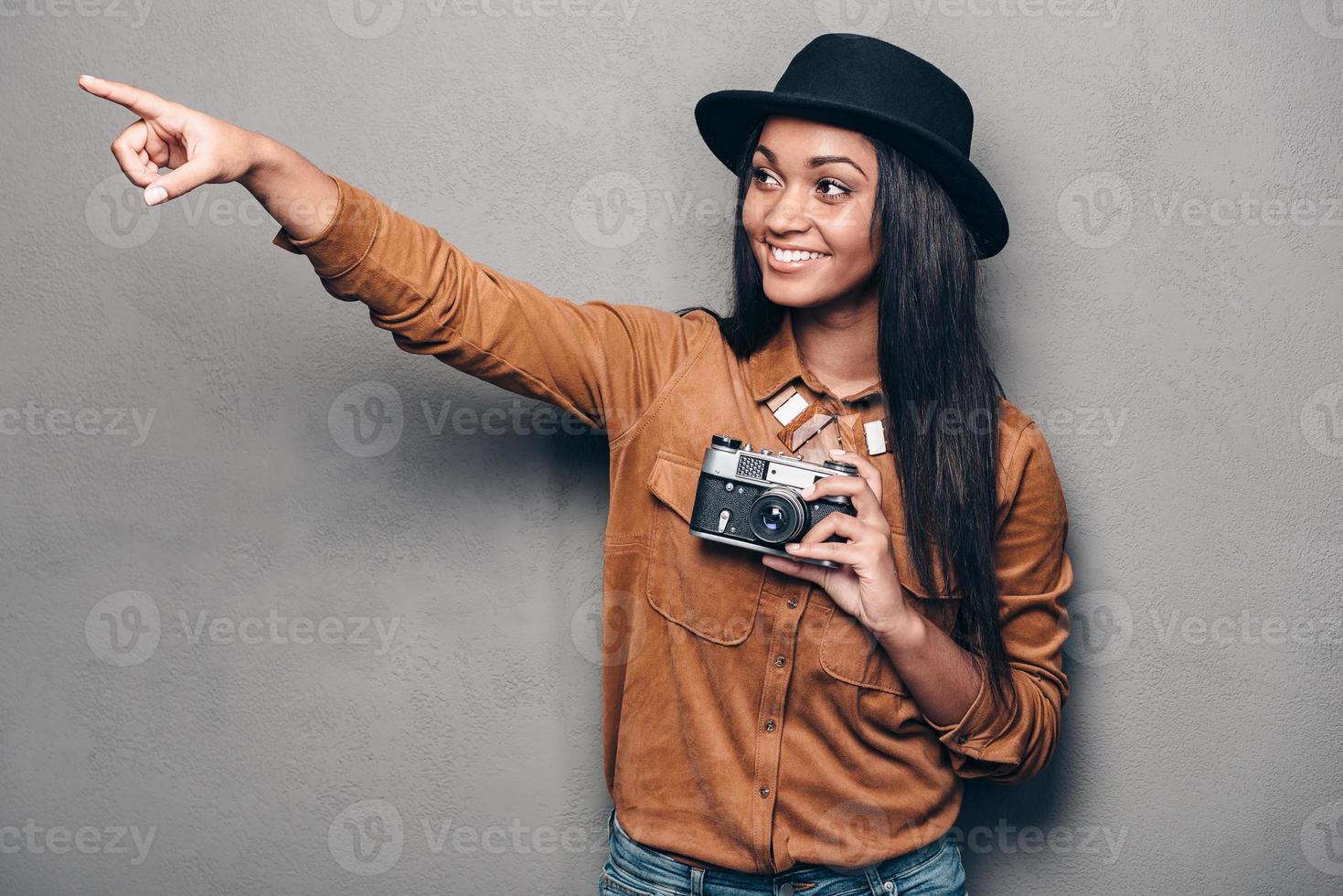 tomemos una foto allí, una hermosa y alegre joven africana sosteniendo una cámara de estilo retro y señalando con una sonrisa mientras se enfrenta a un fondo gris
