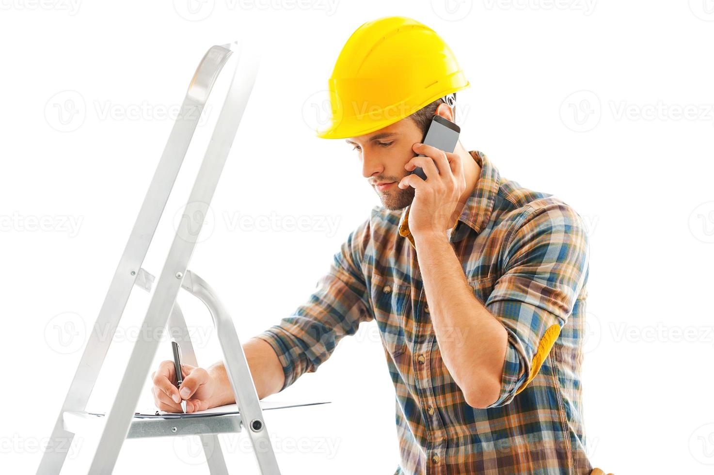 Construction is a serious business. Serious young repairman writing in clipboard and talking on the mobile phone while standing indoors photo