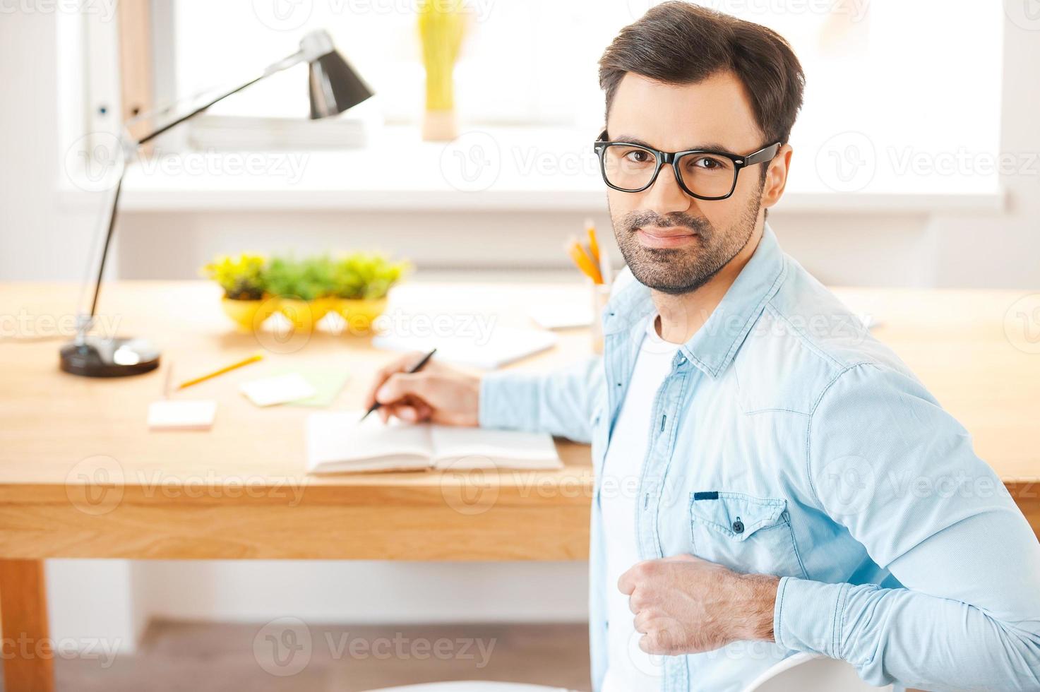 Focused on success. Handsome young man in shirt and eyewear writing in note pad and looking at camera over shoulders while sitting at his working place photo