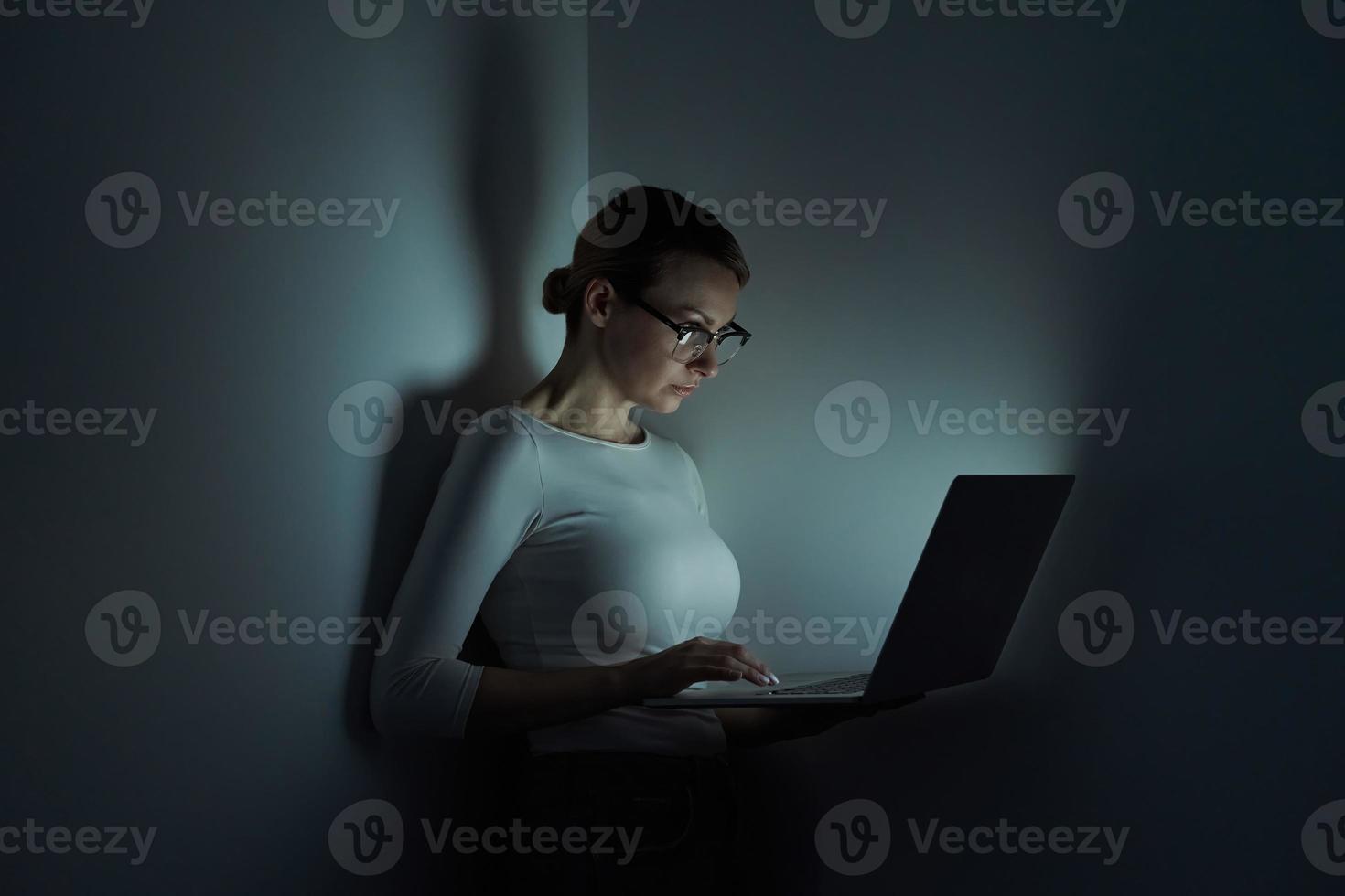 Beautiful young woman working on laptop against a dark wall photo