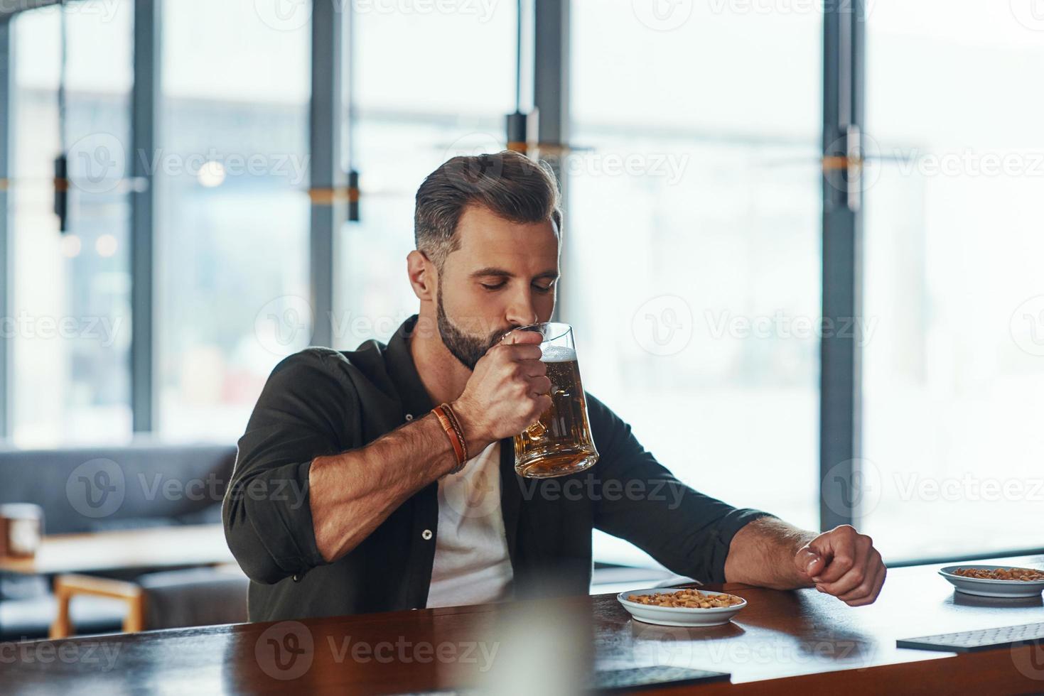 Handsome young man in casual clothing drinking beer while spending time in the pub photo