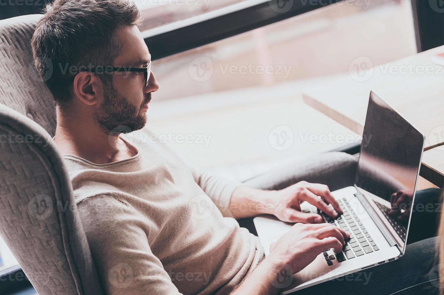 Working with pleasure. Side view of handsome man using his laptop while sitting in chair in front of window photo
