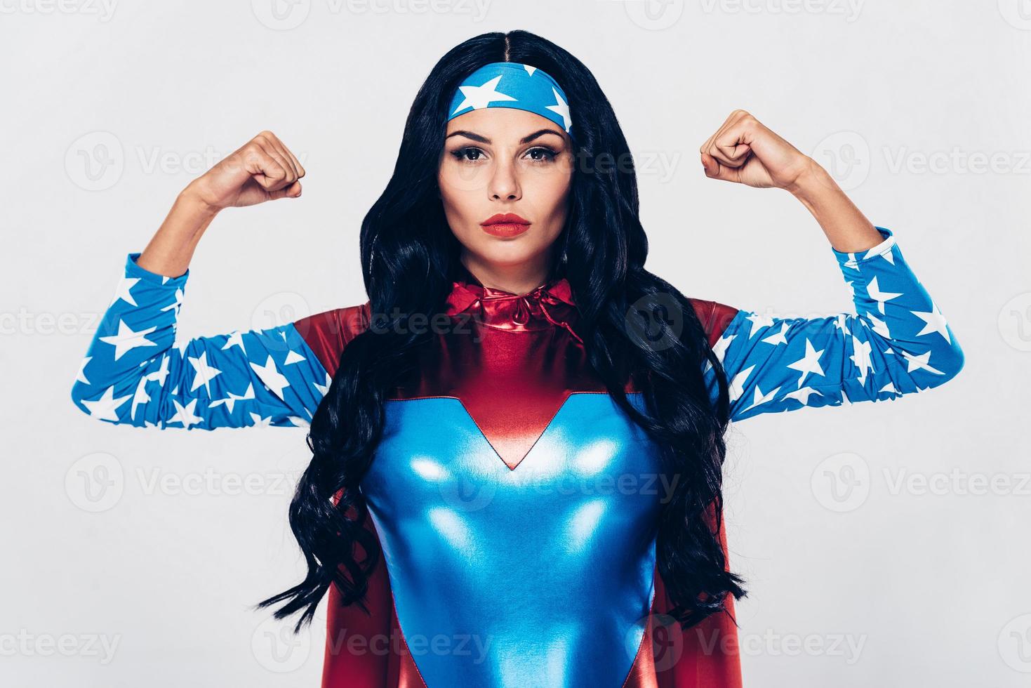 ella tiene mucha fuerza. Hermosa mujer joven en traje de superhéroe mirando a la cámara y flexionando sus bíceps mientras está de pie contra el fondo blanco. foto