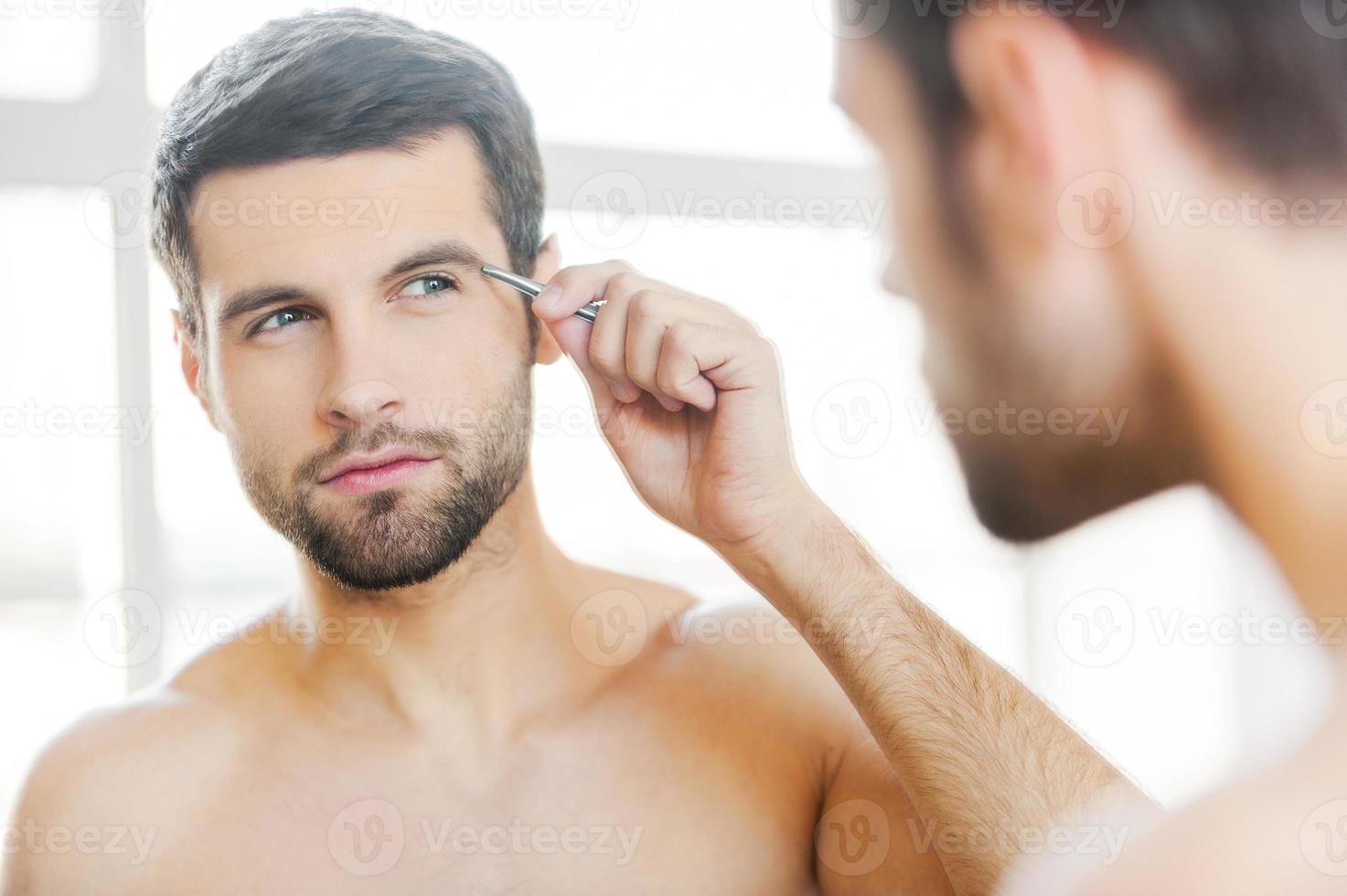 Tweezing his eyebrows. Handsome young man tweezing his eyebrows and looking at himself while standing in front of the mirror photo