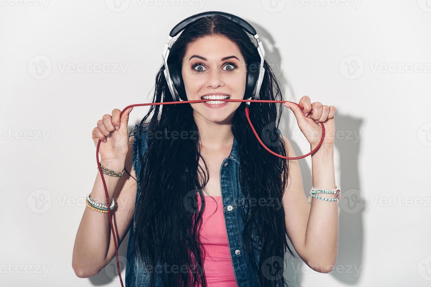 Music lover. Beautiful young woman in headphones carrying wire in mouth and looking excited while standing against grey background photo