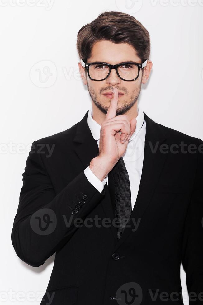 Keeping business secrets. Serious young man in formalwear holding finger on lips and looking at camera while standing against white background photo