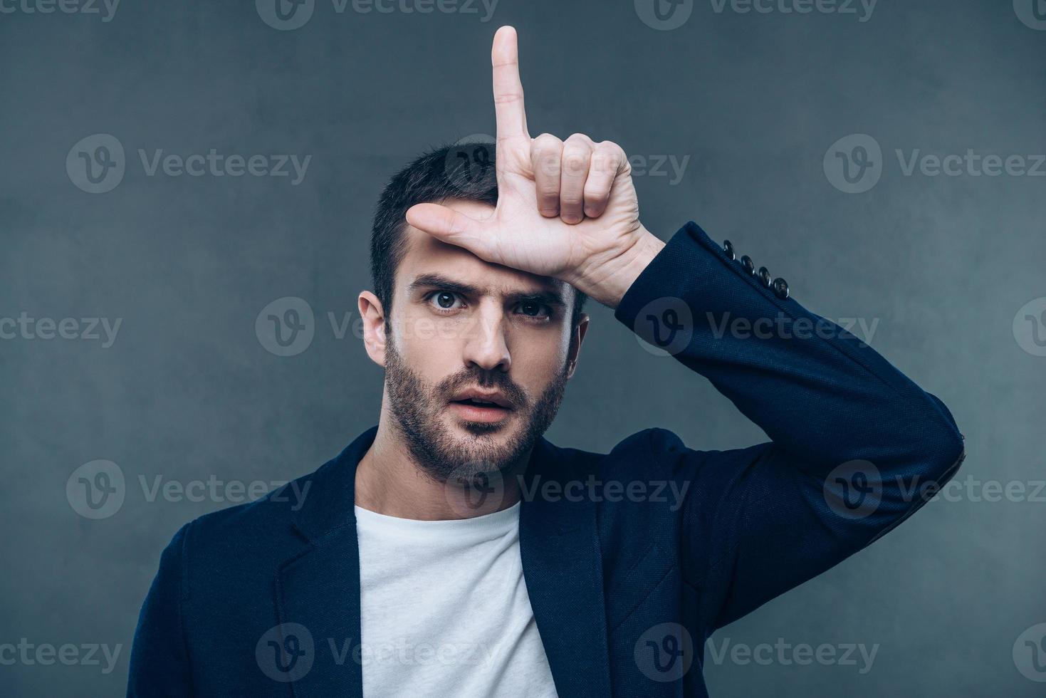 Looser. Portrait of beautiful young man gesturing L sign while standing against grey background photo