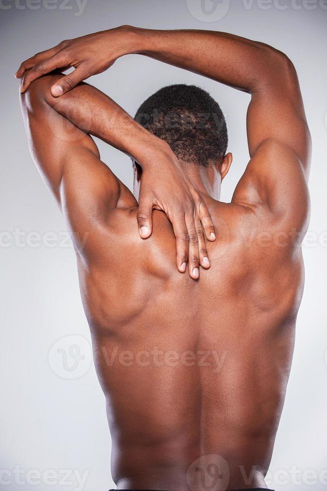 dolor en las articulaciones. vista trasera de un joven africano sin camisa tocando su cuello y codo mientras está de pie contra un fondo gris foto