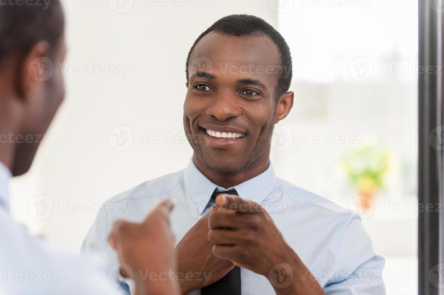 Feeling confident. Young African man adjusting his necktie and pointing himself at the mirror photo