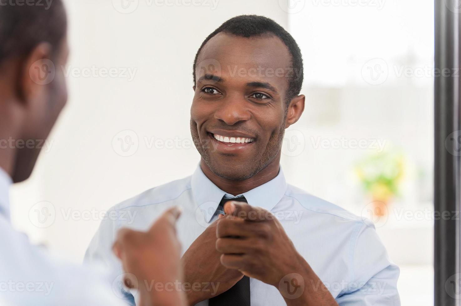 con mucha confianza. joven africano ajustándose la corbata y apuntándose al espejo foto