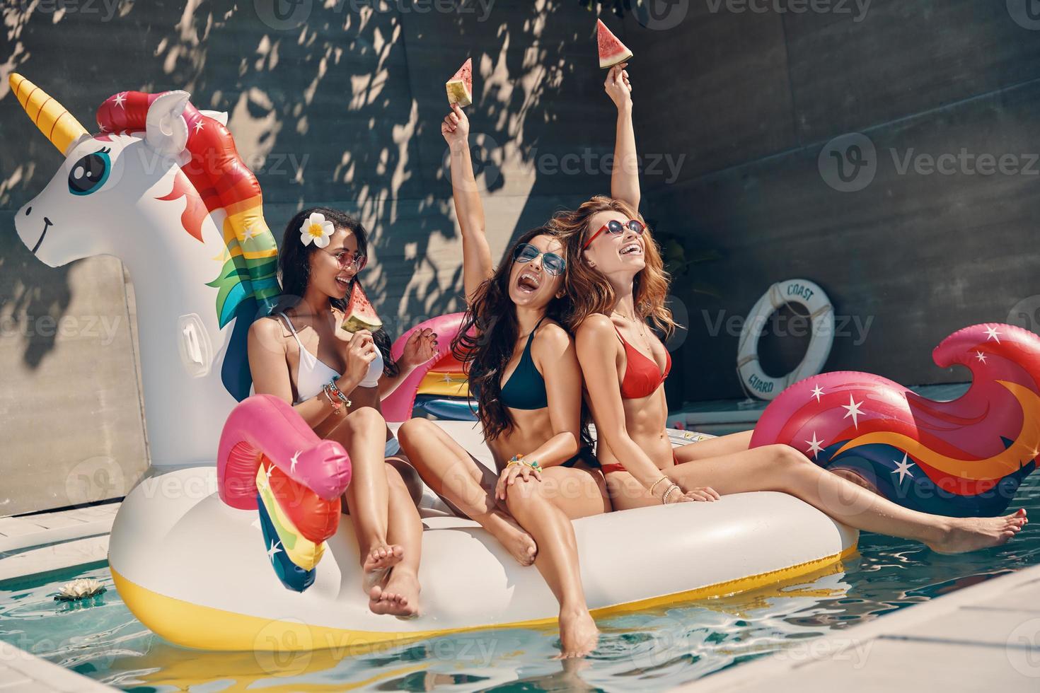 Attractive young women in swimwear smiling and eating watermelon while floating on inflatable unicorn in swimming pool outdoors photo