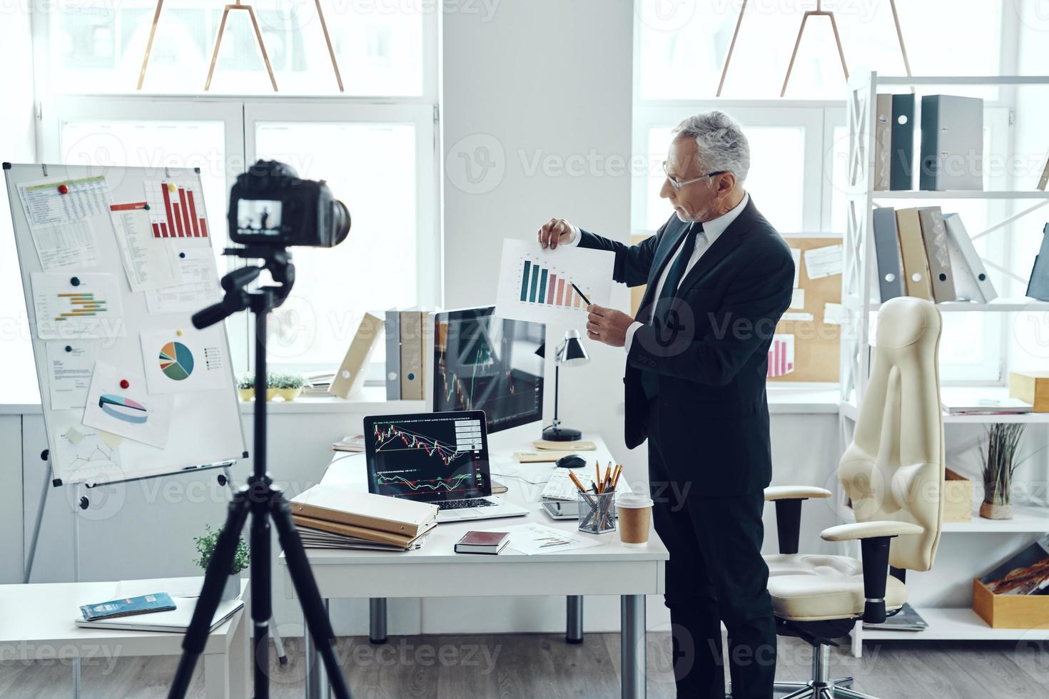 Senior man in elegant business suit pointing at chart while making social media video photo