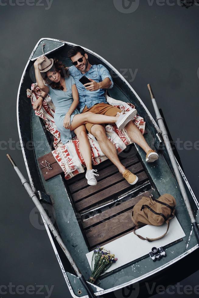 Great time together. Top view of beautiful young couple taking selfie using smart phone while lying in the boat photo