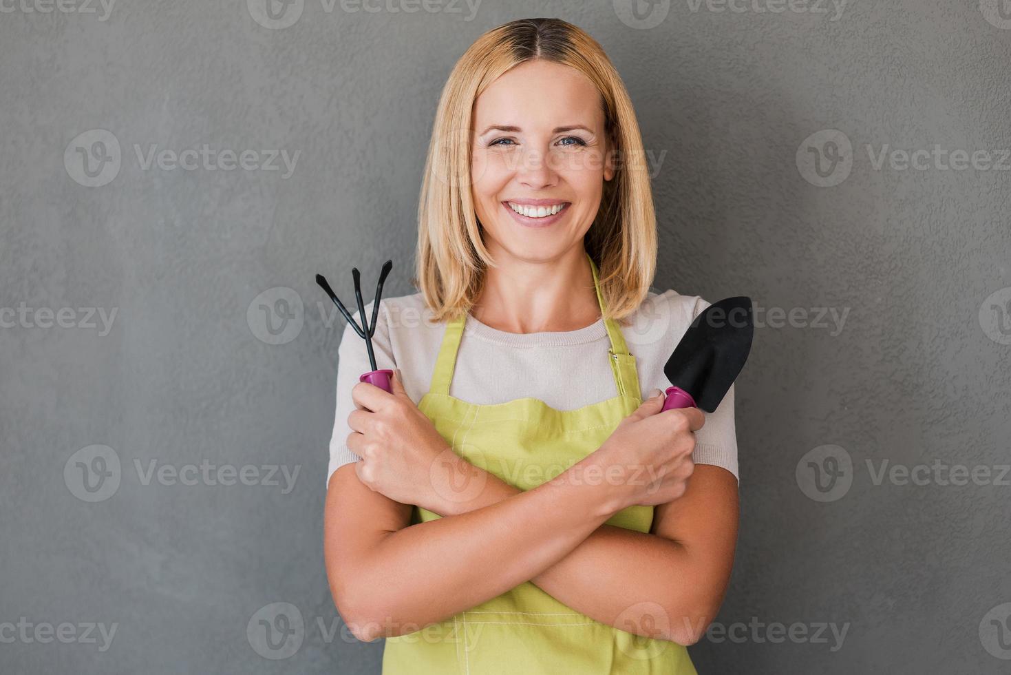 Ready for gardening. Happy mature woman in green apron holding gardening equipment and smiling while standing against grey background photo