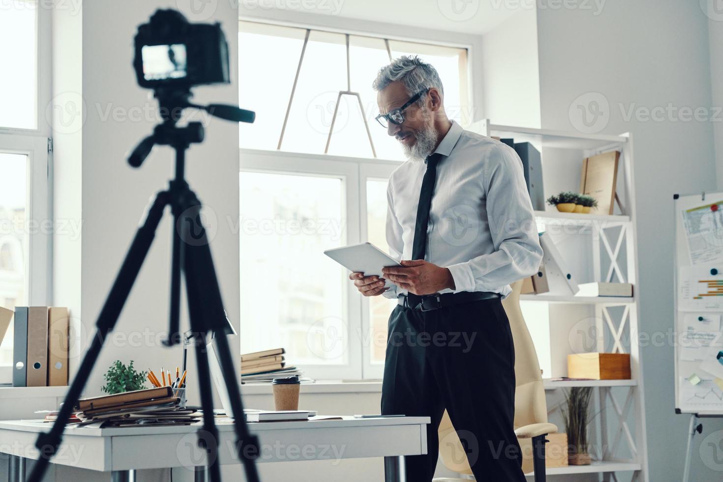 Confident mature man in elegant shirt and tie using digital tablet while making social media video photo