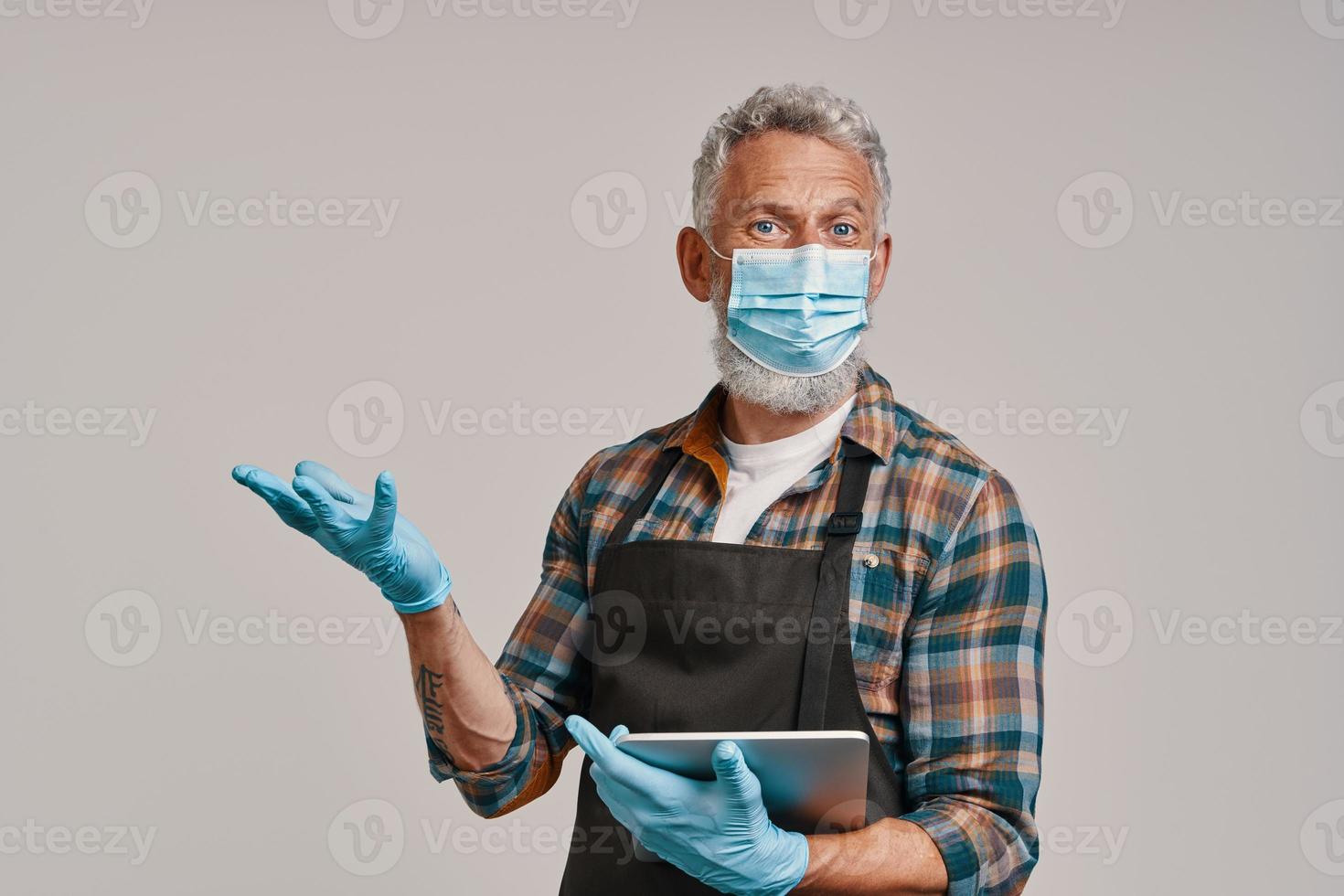 Senior man in apron and protective face mask holding digital tablet and gesturing photo