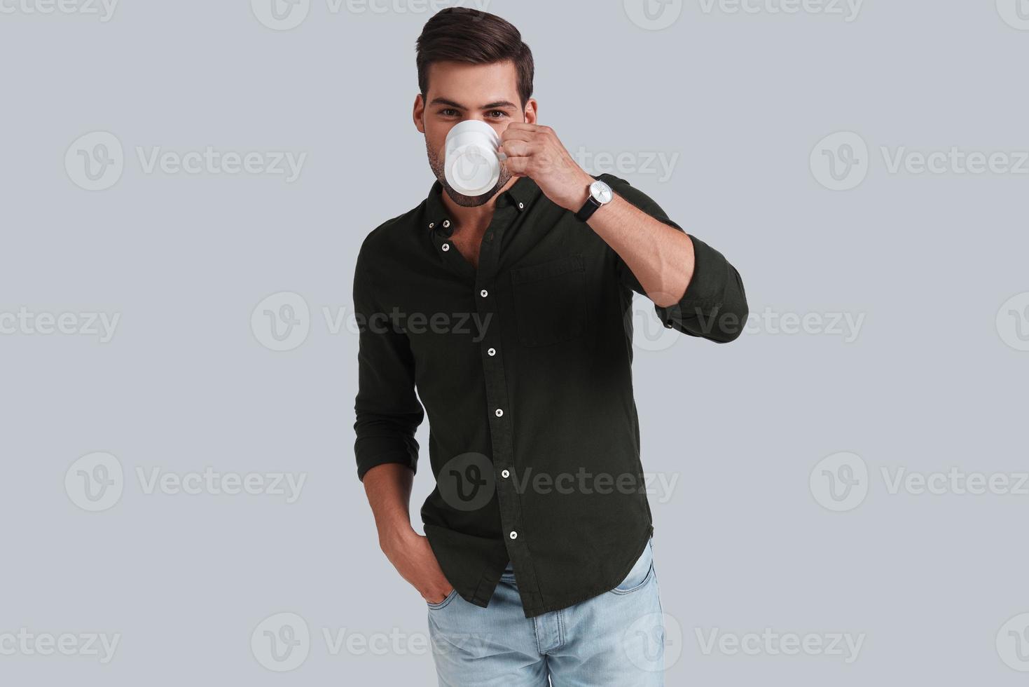 Little coffee break.  Good looking young man drinking while standing against grey background photo