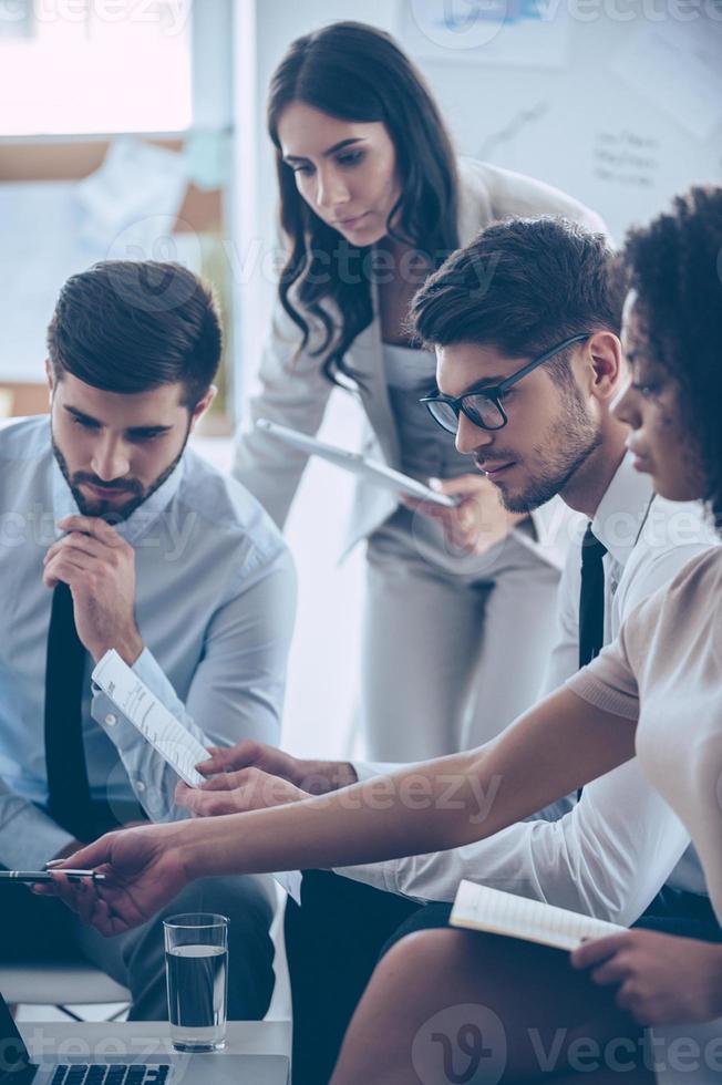 Take a look at this numbers Young beautiful African woman pointing at her laptop while sitting on the couch at office with her coworkers photo