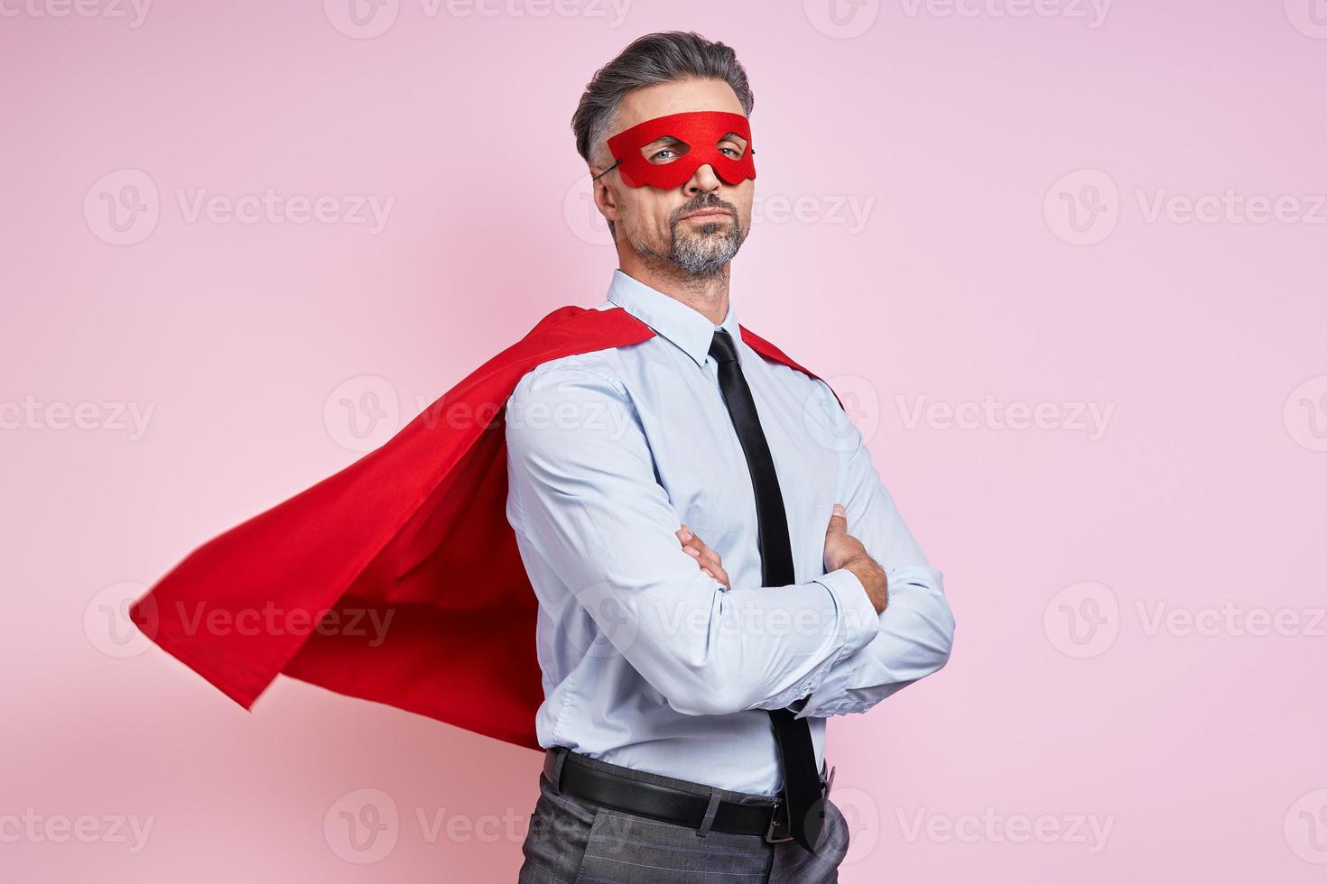 hombre confiado en camisa y corbata con capa de superhéroe y manteniendo los brazos cruzados contra el fondo rosa foto